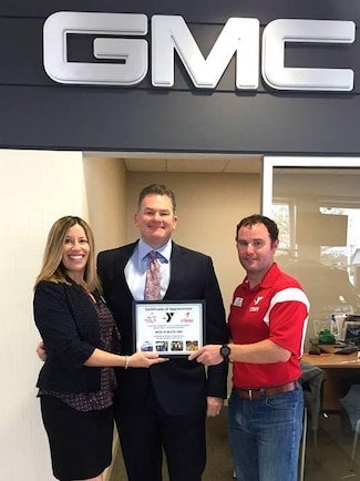 Three people holding a certificate of appreciation under a large GMC logo